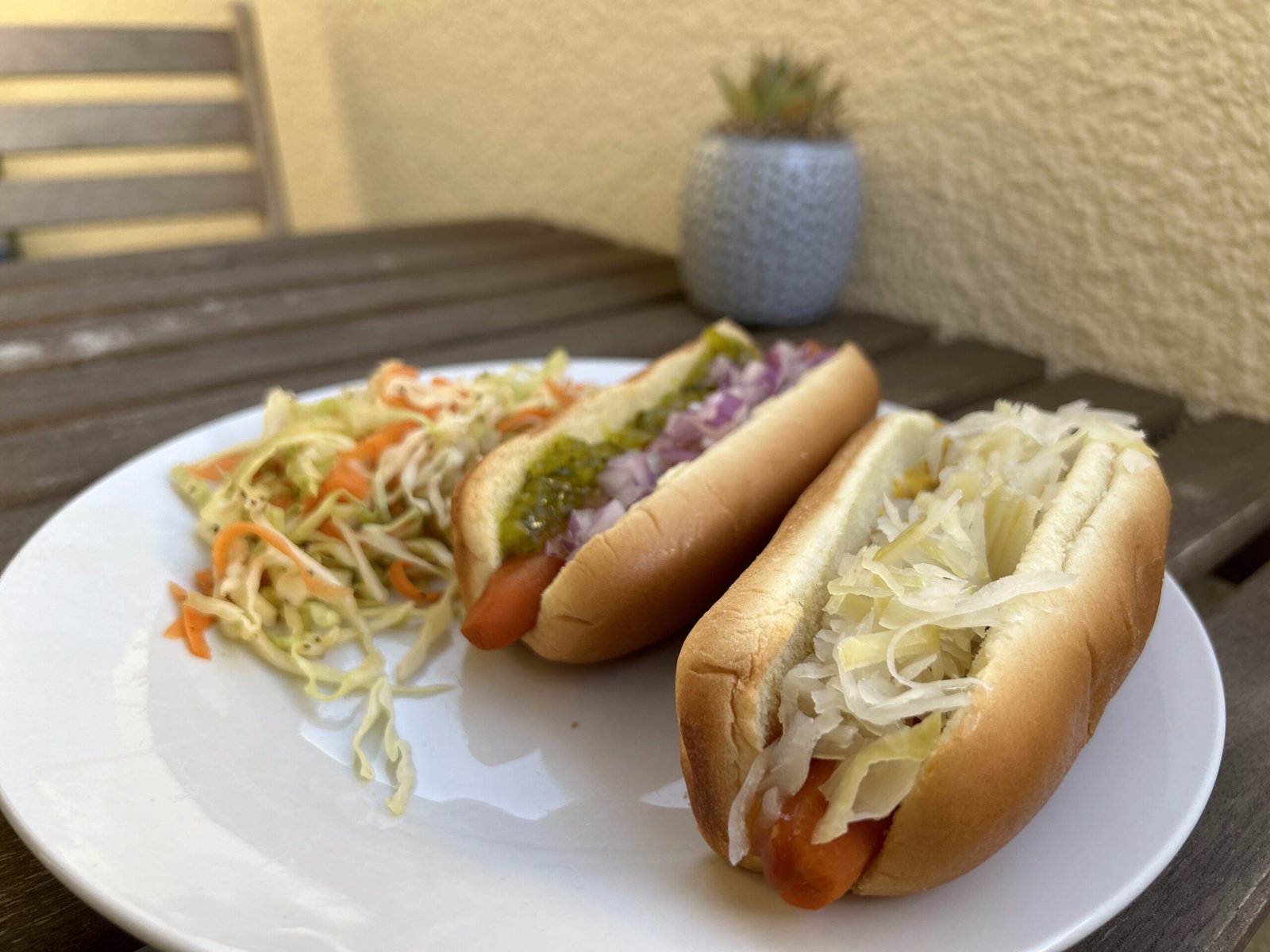 Two carrot hotdogs with sauerkraut and coleslaw on a white plate on an outdoor wooden table next to a succulent